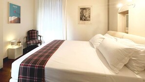 A hotel room with a bed, a chair, a nightstand, a window with curtains, and wall-mounted lights. - COMO mi CASA • Lakefront • Ideal Base for Visiting the Lake (Malgrate)