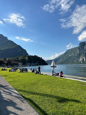 Ein grasbewachsenes Ufer mit Menschen, die entspannen, einem Segelboot und Bergen im Hintergrund.