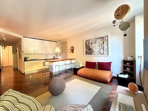 Ein modernes Wohnzimmer mit einer Couch, einem runden Hocker, einem Essbereich mit Bar und einer Küche im Hintergrund.