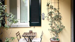 A patio with a table and chairs, a window with shutters, a potted plant, and a wall-mounted plant holder. - COMO mi CASA • Lakefront • Ideal Base for Visiting the Lake (Malgrate)