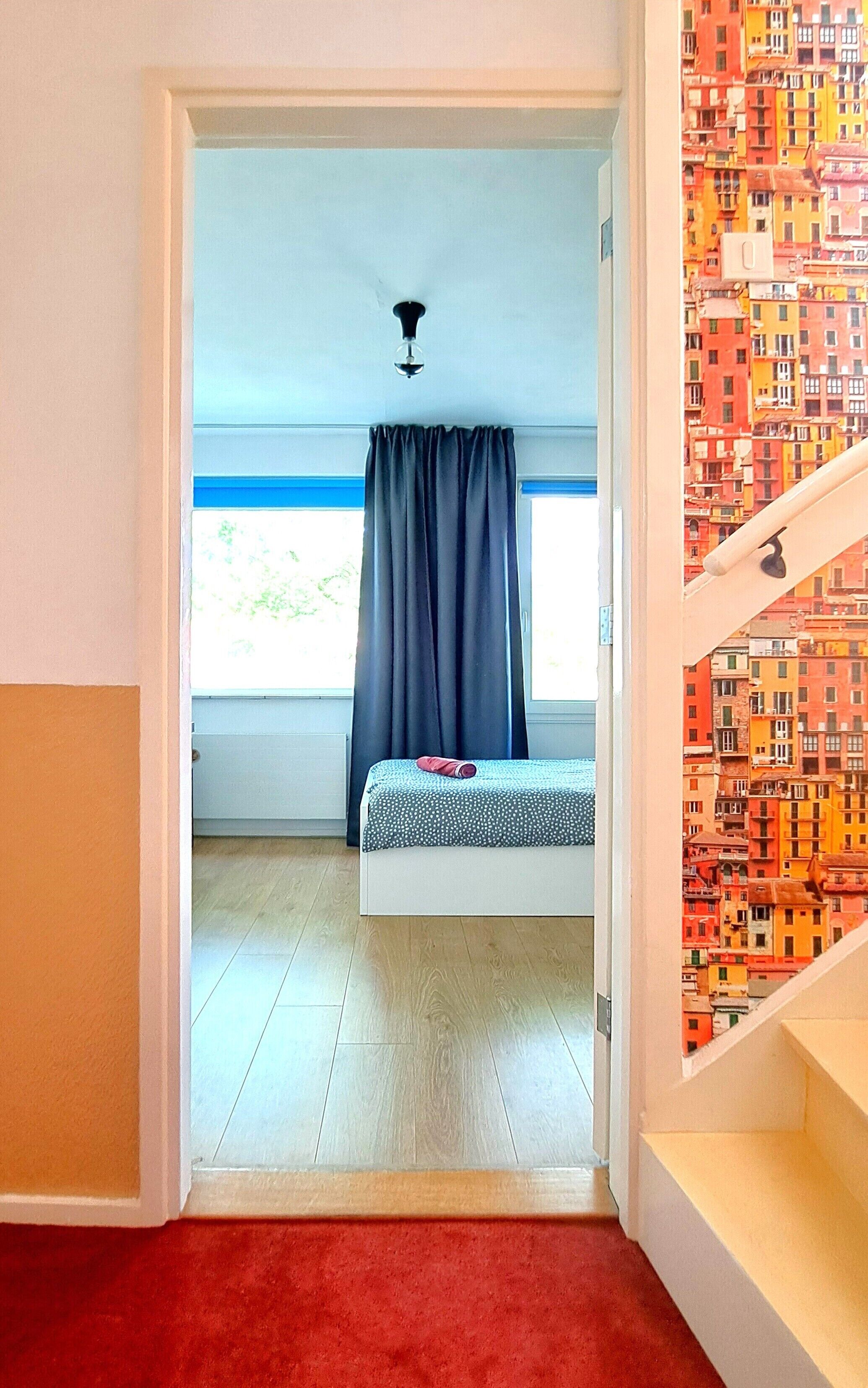 Une chambre à coucher avec un lit, une fenêtre avec des rideaux, un luminaire au plafond et une vue sur des bâtiments aux couleurs vives par la fenêtre.