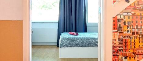 Une chambre à coucher avec un lit, une fenêtre avec des rideaux, un luminaire au plafond et une vue sur des bâtiments aux couleurs vives par la fenêtre.