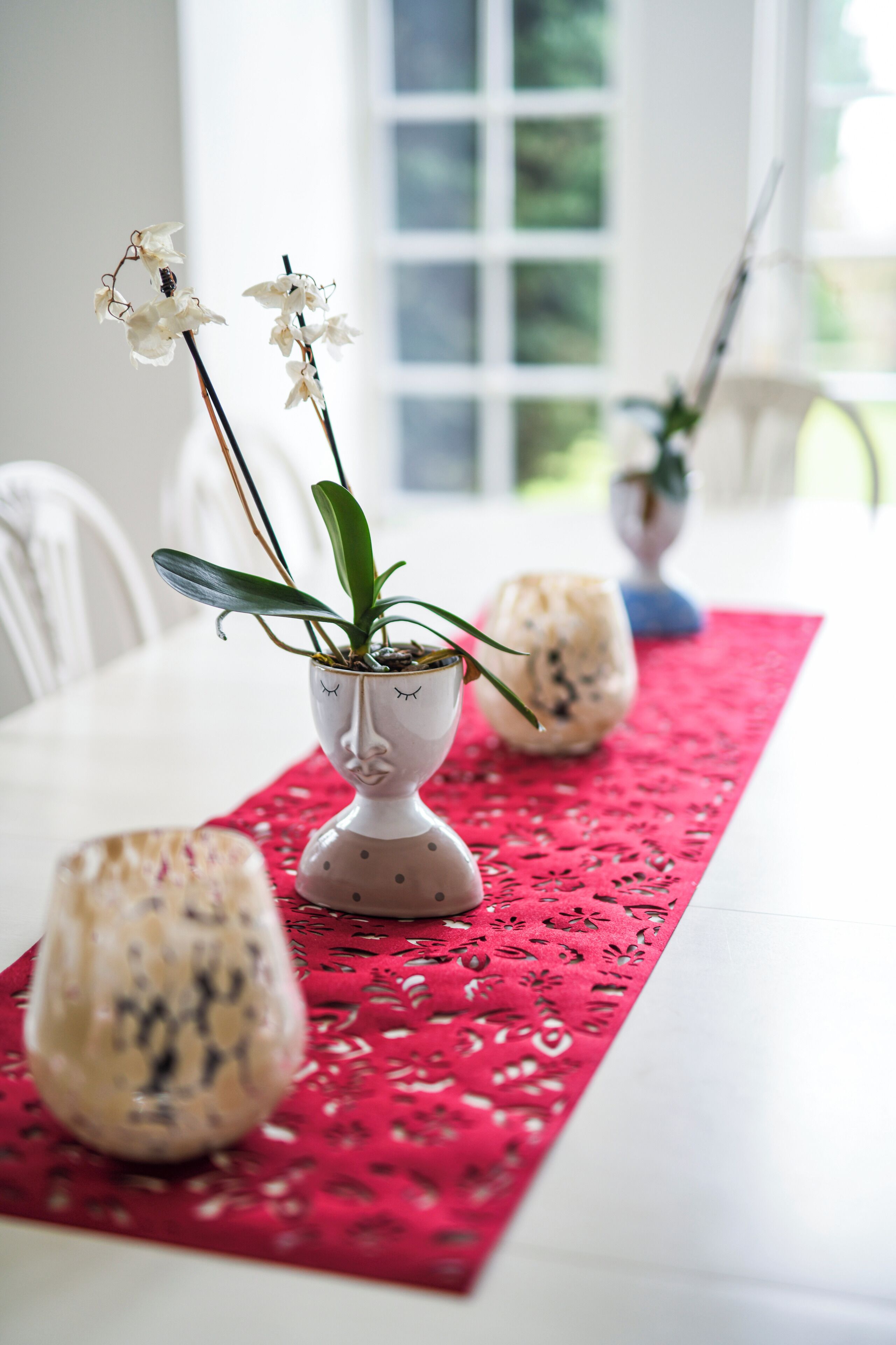 A dining table with a red table runner, two white ceramic vases with orchids, and a decorative glass on the table.
