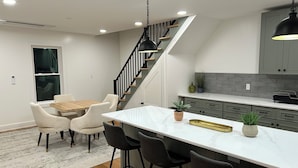 A modern kitchen with a central island, bar stools, a dining table with chairs, and a staircase with a black railing.