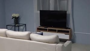 Un salon avec un canapé blanc, un téléviseur à écran plat posé sur un meuble en bois, une petite table avec un vase et une fenêtre ornée de rideaux légers.