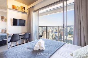 Habitación de hotel con cama, escritorio, dos sillas, televisor y una ventana grande con vistas a la ciudad.