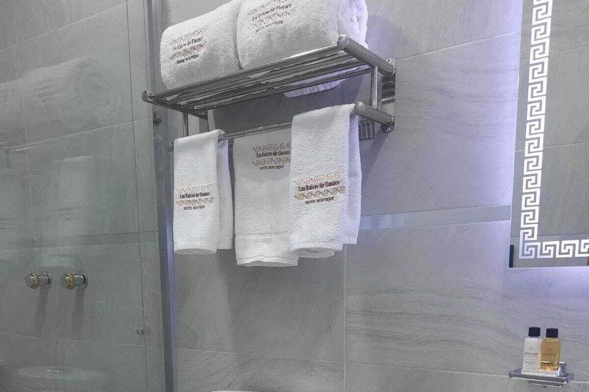 Un baño con inodoro, ducha, lavamanos y un cubo de basura. Hay toallas colgadas en un perchero sobre el inodoro.