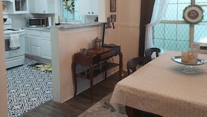 Une pièce comprenant une table à manger, un lustre, un coin cuisine avec des meubles blancs et un tapis à motifs.
