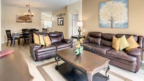 A living room with a brown leather sofa, a coffee table, and a ceiling fan. There is a dining area in the background with a table and chairs. - Resort-Style Villa | Pool, Spa & Game Room! (Kissimmee)