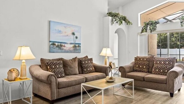 A living room with two sofas, a coffee table, and two lamps. There is a painting on the wall and a window with plants. - Sunny Private Pool & Spa & Fenced Yard. Nr Parks (Kissimmee)