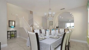 Un espace repas avec une longue table dressée pour six personnes, des chaises blanches et un lustre.