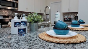 Una cocina con encimeras de granito, armarios blancos, microondas de acero inoxidable, cafetera y cuencos de cerámica azul sobre caminos de tela.