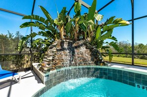 A swimming pool with a waterfall feature, surrounded by large banana plants and a stone wall. - Spacious 7Br Villa In Kissimmee Up To 14 Guests (Kissimmee)