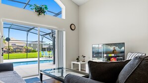 A living room with a glass door leading to a pool, a flat-screen TV, and a clock on the wall. - Emerald Island Retreat Near Parks (Kissimmee)