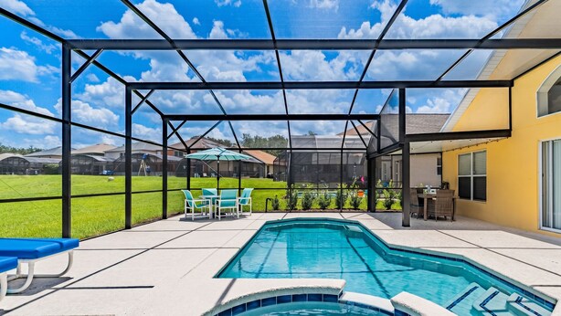 A covered outdoor swimming pool with a view of a green lawn and a yellow building. - Emerald Island Retreat Near Parks (Kissimmee)