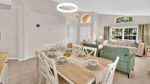 Un coin repas avec une table en bois dressée pour quatre personnes, des chaises blanches et un canapé vert en arrière-plan.