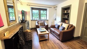 A living room with a fireplace, a sofa, a coffee table, and a bookshelf. - Gate Lodge - historic rural cottage with hot tub, Aberdeenshire (Fettercairn)
