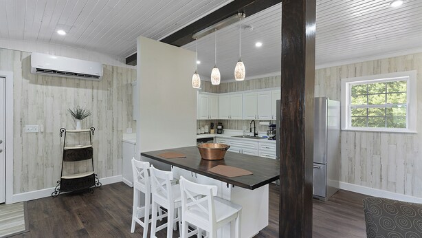 A modern kitchen with a central island, white bar stools, pendant lights, and a view of the outdoors through a window. - Beyond Charming Luxury Barndominium - Get away without going far away! (Howey-in-the-Hills)