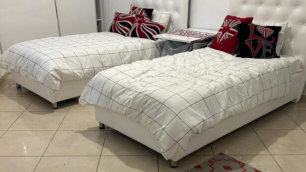 Un dormitorio con dos camas, cada una con cabeceros blancos acolchados y ropa de cama a juego. Una alfombra estampada se encuentra frente a las camas.