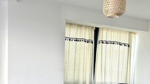 Un sofá seccional gris con varios cojines, una lámpara tejida y una ventana con cortinas translúcidas.