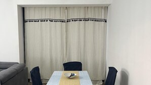Una habitación pequeña y bien iluminada con una mesa de comedor lista para dos personas, rodeada de sillas. Un sofá se encuentra a la izquierda, y una ventana con cortinas se ve al fondo.