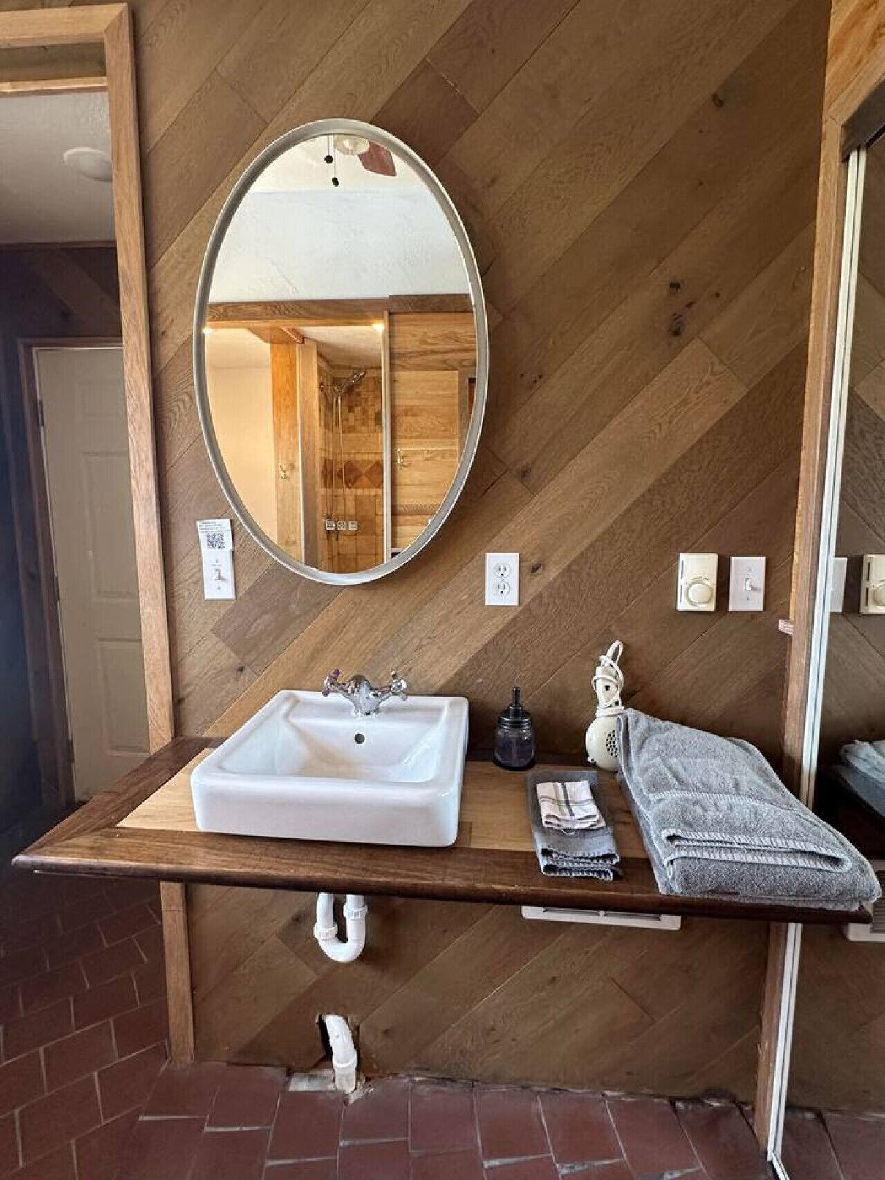 Une salle de bain avec un meuble-lavabo en bois, un miroir rond, un lavabo blanc, une serviette et une douche visible dans le reflet.