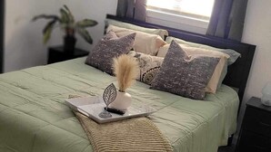 A neatly arranged bedroom with a bed, a bedside table, a nightstand, a window with curtains, and a framed picture on the wall. - The Hideaway (Sacramento)