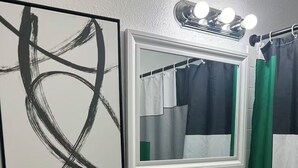 A bathroom with a toilet, sink, and mirror. There is a black and white abstract painting on the wall, a potted plant, and a shower curtain. - The Hideaway (Sacramento)