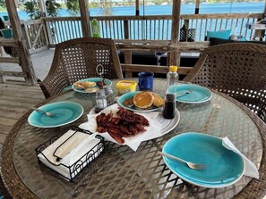 Uma mesa redonda de vidro com cadeiras de vime, com pratos de panquecas, bacon e uma bebida.
