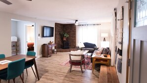 A living room with a wooden floor, a brick accent wall, a fireplace, a TV, and a dining area with chairs and a table.