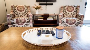 A living room with two patterned armchairs, a coffee table with remote controls, a candle, and a TV on the wall.