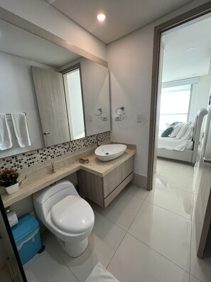 Une salle de bain moderne avec des toilettes blanches, un lavabo beige et un grand miroir. On y trouve une poubelle bleue et une petite plante posée sur le comptoir.