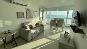 Een moderne woonkamer met een bank, een eettafel met stoelen, een aan de muur bevestigde televisie en een groot raam met uitzicht op de zee.