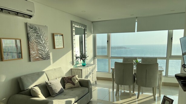 Een moderne woonkamer met een bank, een eettafel en stoelen. Er is een groot raam met uitzicht op zee.