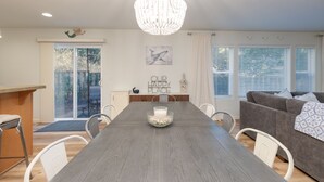 A modern dining area with a wooden table, white chairs, a chandelier, and a glass door leading outside. - OMalley's Ocean Getaway—Nelscott Bch-Dog OK-EV (Lincoln City)