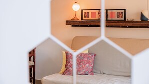 A cozy room with a wooden shelf displaying framed pictures and a lamp, a grey sofa with patterned cushions, and a white geometric wall decoration.