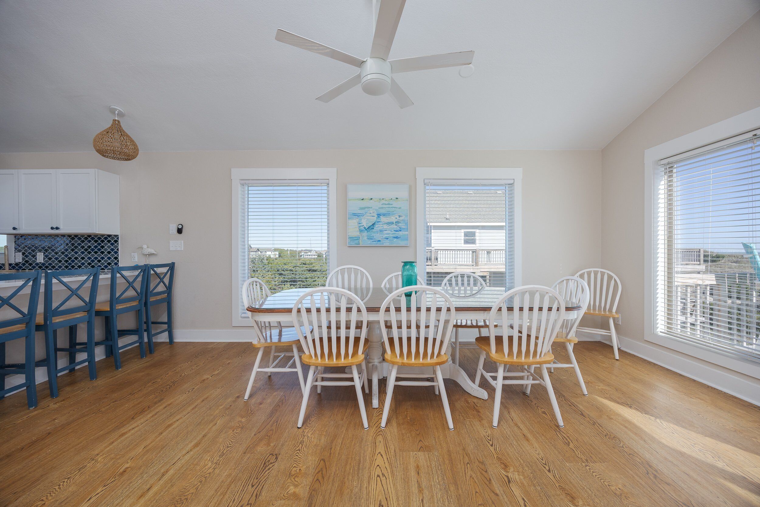 A dining area with a white table and chairs, a ceiling fan, and large windows with blinds.