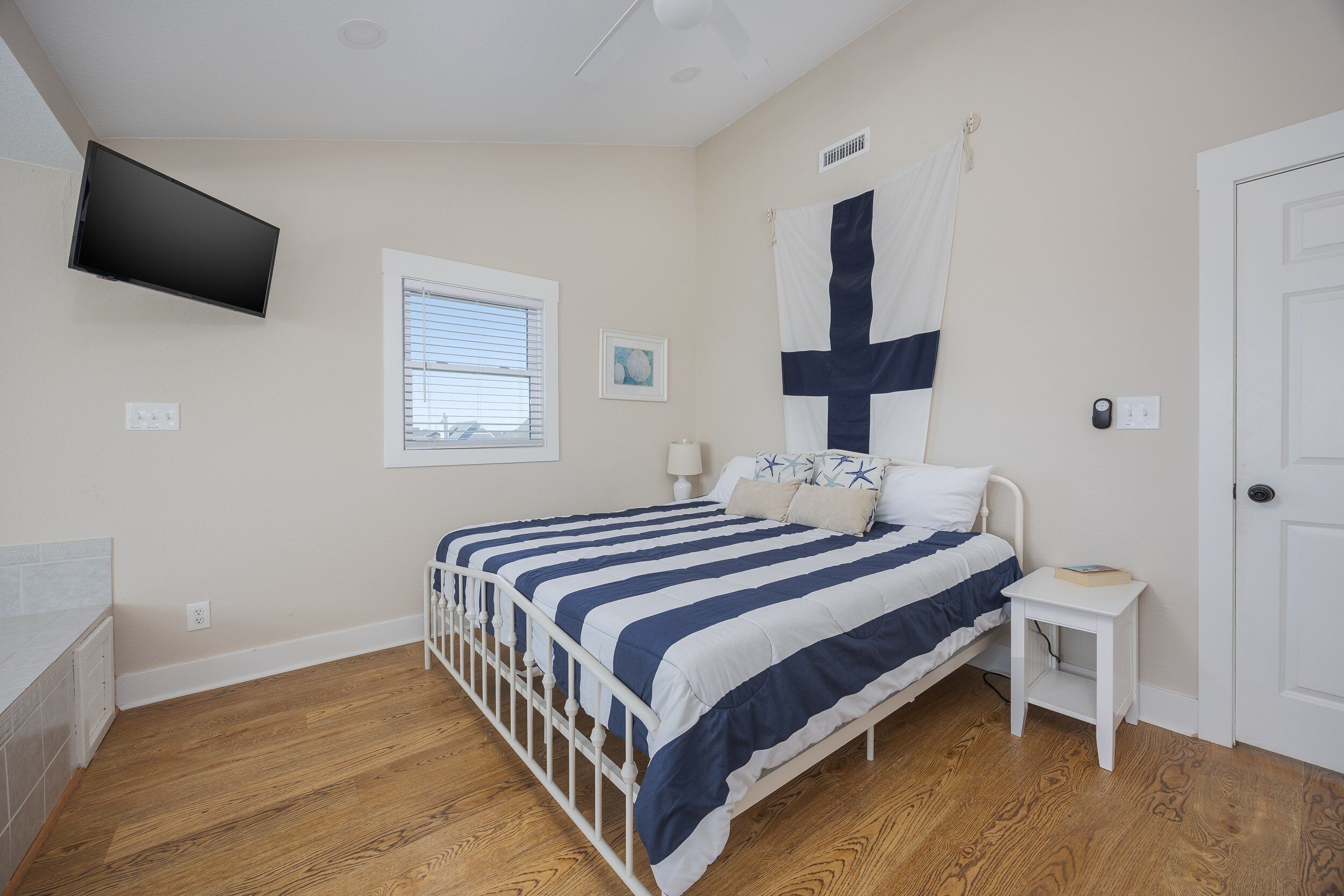 A bedroom with a bed, a nightstand, a TV mounted on the wall, a window with blinds, and a door.