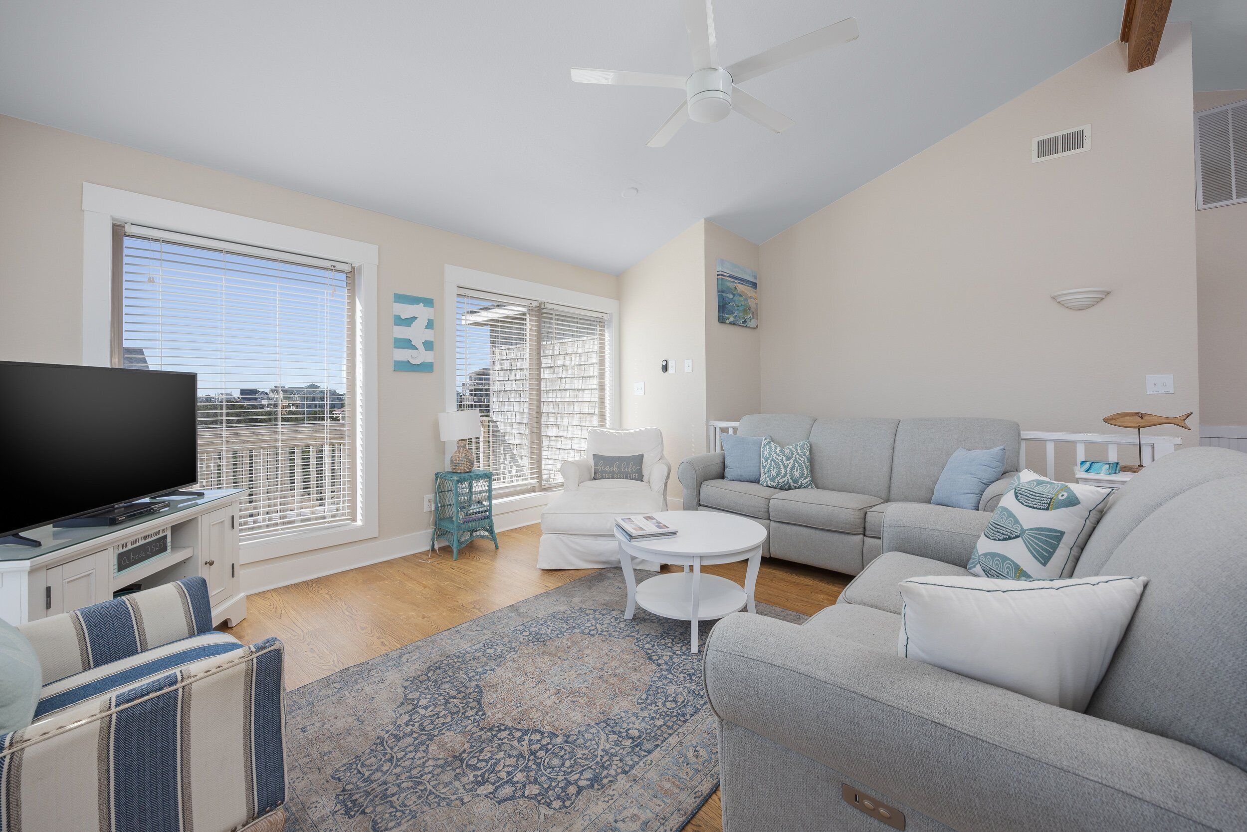 A living room with a flat-screen TV, a grey sofa, a white armchair, a patterned rug, and a ceiling fan.