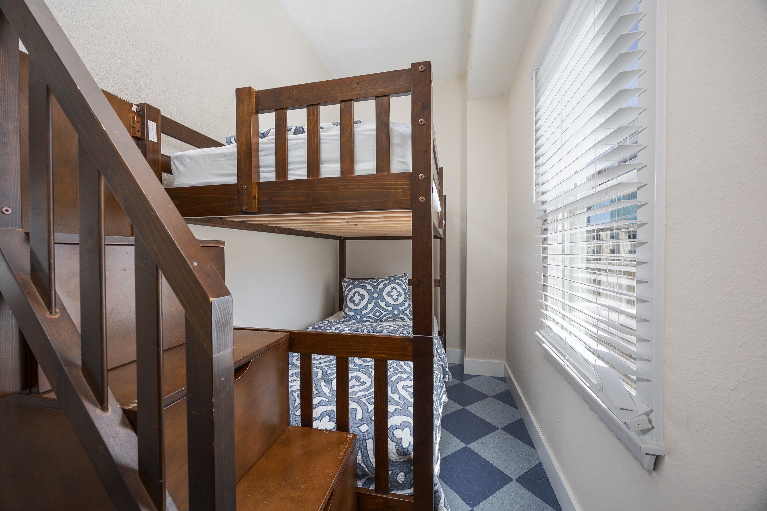 Un lit superposé avec une structure en bois et une couvre-lit à motifs, un escalier en bois et une fenêtre avec des stores.
