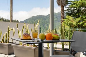 Une table avec un croissant, des fruits et des boissons sur un balcon agrémenté de plantes en pot, offrant une vue sur la montagne.