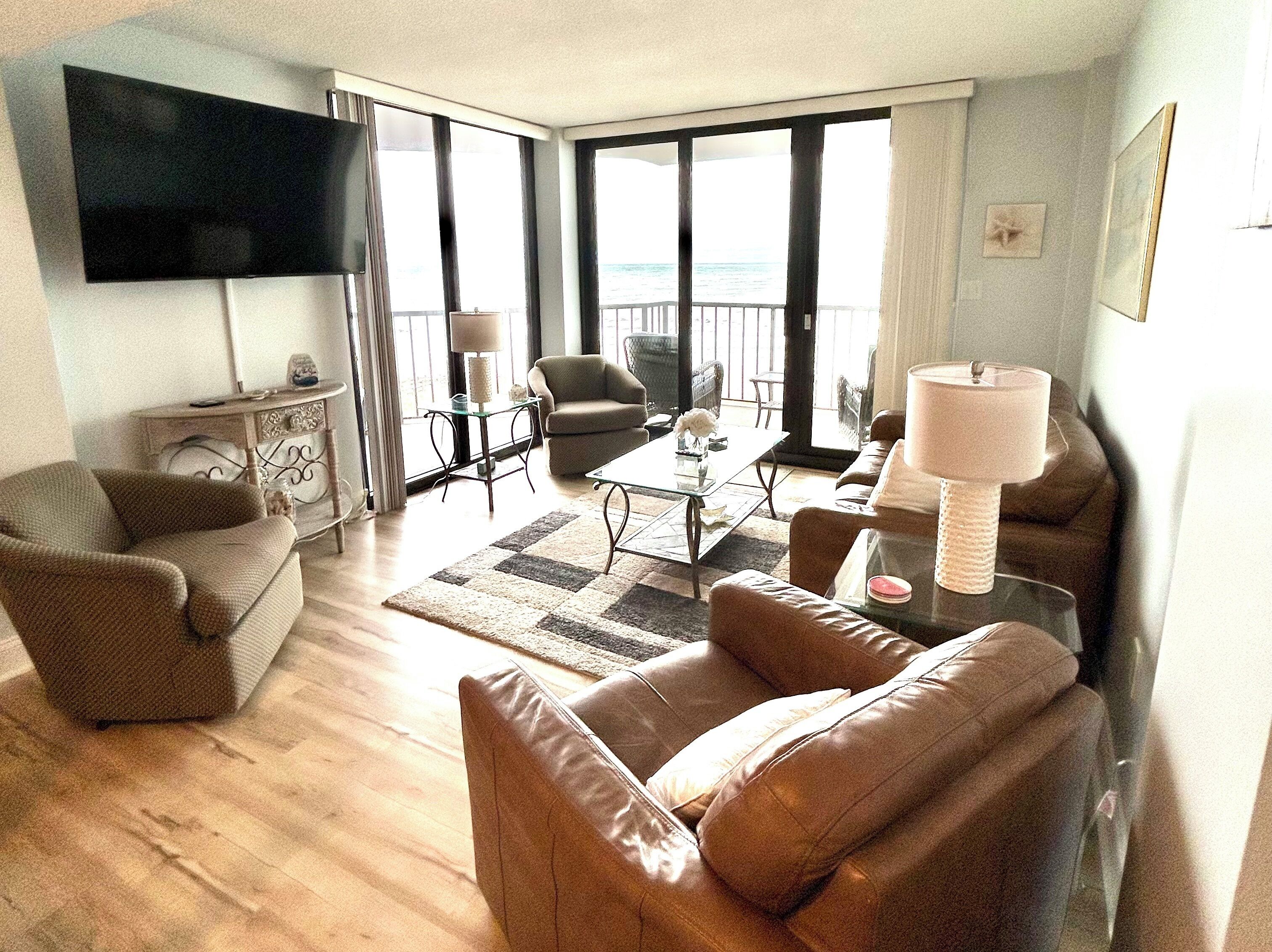 A living room with a flat-screen TV, leather sofas, a glass coffee table, and a view of the sea through large windows.