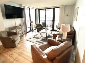 A living room with a flat-screen TV, leather sofas, a glass coffee table, and a view of the sea through large windows.