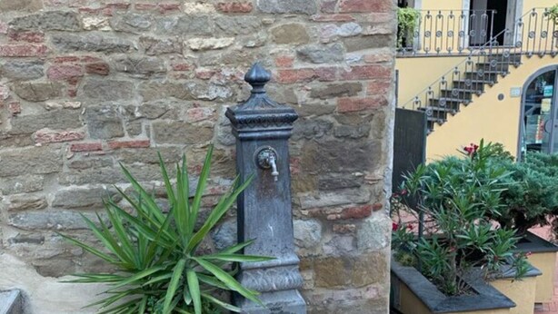 Een stenen fontein met een tuit, omringd door plantenpotten, voor een stenen muur en een geel gebouw met een trap.
