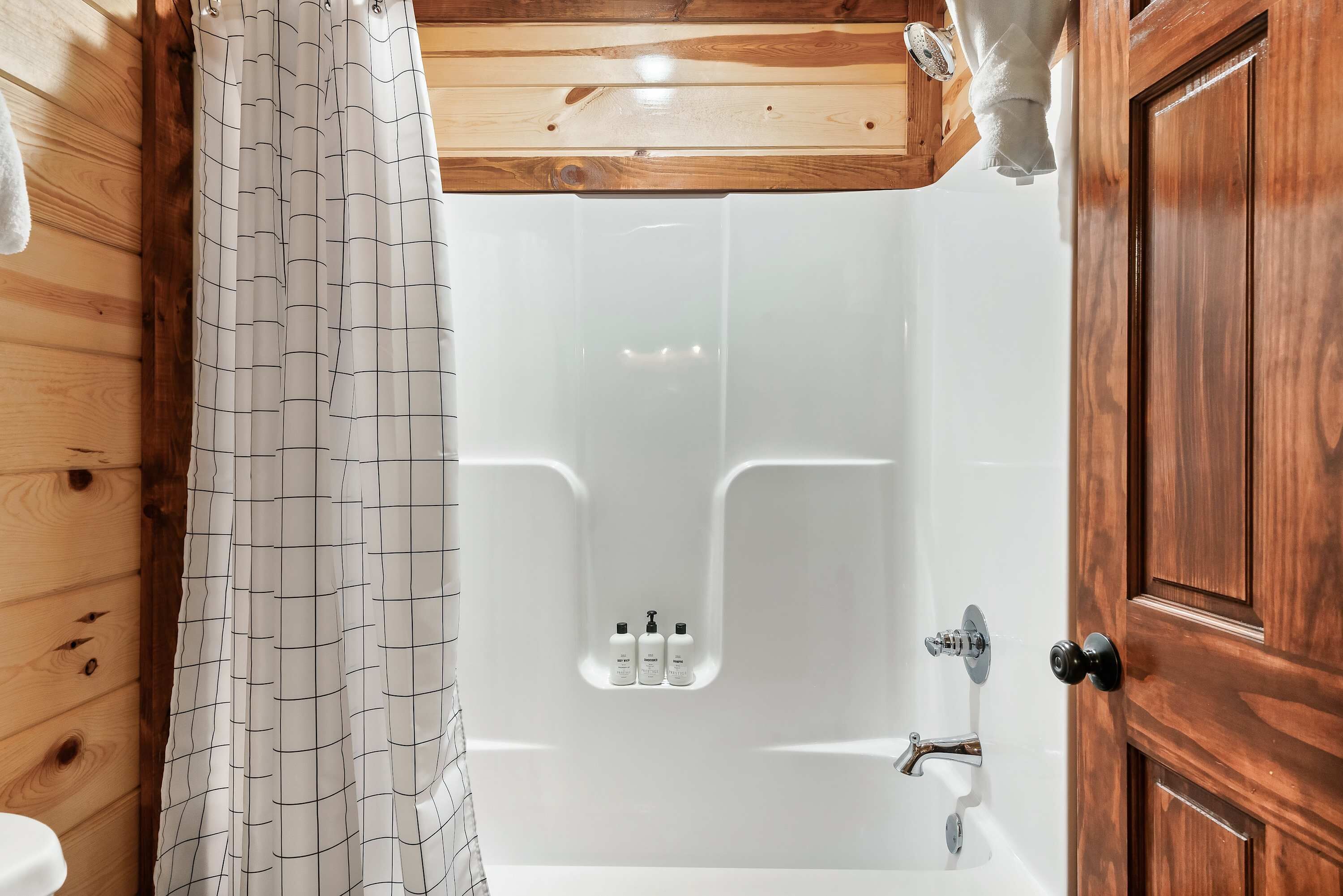 Un espace douche avec une structure en bois, un mur recouvert de carreaux blancs, une porte en bois et un rideau de douche à motifs géométriques.