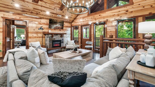 A log cabin interior with a fireplace, wooden furniture, and large windows.