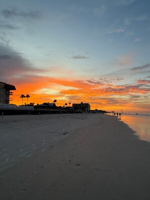 A beach with a sunset, buildings, and palm trees.