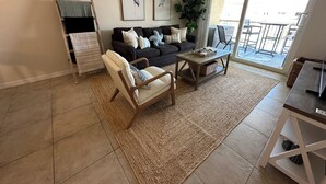 A living room with a sofa, armchair, coffee table, and a large window with a view of a balcony. - Top Floor Remodeled 2BR/2BA Beachfront Condo (New Smyrna Beach)