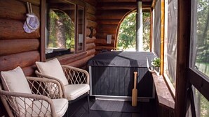 Una cabaña de madera con un porche de madera, dos sillones de mimbre, una zona de estar al aire libre techada y vistas al bosque circundante.