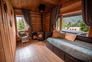 A log cabin interior with a bed, a chair, a wood-burning stove, and a view of the outdoors. - Off-Grid Wilderness Cabin with Mountain Views, Hot Tub, Sauna & Canoes (Provincial Park)